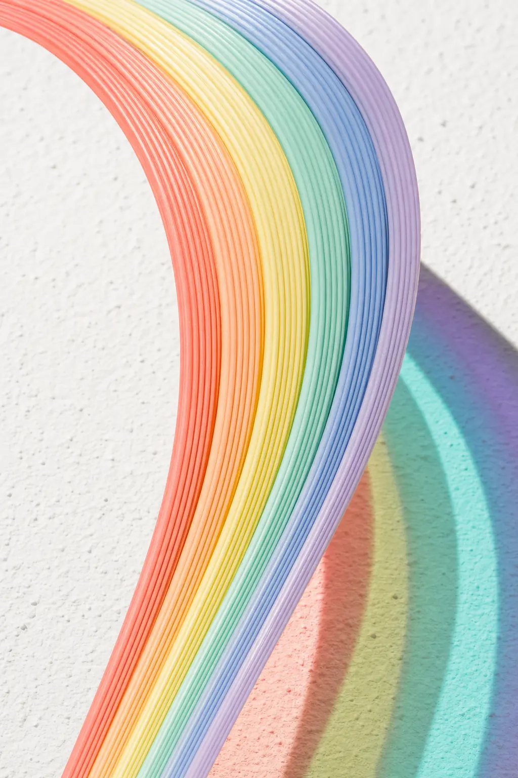 A sweeping bundle of pastel-coloured cable curving across white plaster in hard sunlight, casting a matching band of coloured shadow.